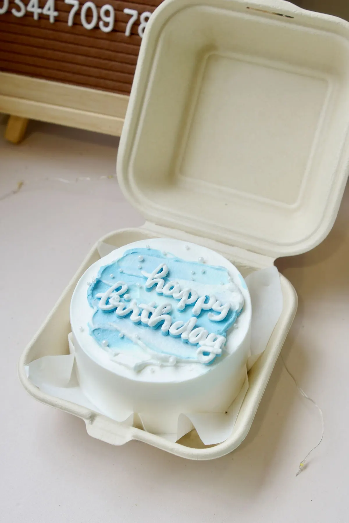 Small white bento cake piped with happy birthday in blue, in an open takeout box